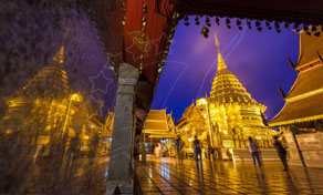 Golden chedi and temple pavilions lit at twilight, reflected on wet tiles, with blurred visitors walking past