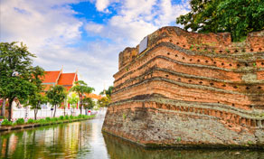 Weathered red brick corner bastion of Chiang Mai's old city wall beside the moat, with temple roofs and palms behind