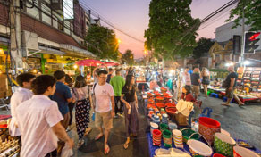 Crowded Thai night market street at dusk with stalls selling plastic buckets and goods under string lights