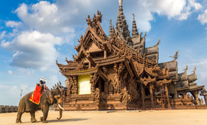 Ornate carved wooden temple in Thailand with an elephant in a red saddle and rider on the sandy foreground under blue sky