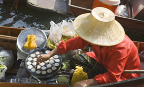 Vendor in a conical straw hat cooks coconut pancakes on a wooden boat at a Thai floating market canal