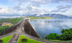 Road along a large earthen dam crest beside a reservoir, with limestone hills and tropical forest under blue sky