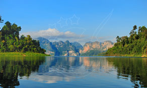 Calm emerald lake reflecting limestone karst peaks and forested shores under a blue sky with white clouds