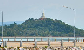 Golden hilltop pagoda on a forested hill seen beyond a concrete road guardrail and rocky embankment