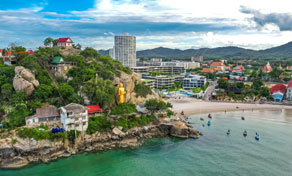Aerial view of a rocky headland with golden Buddha statue above a beach, hotels and fishing boats in a turquoise bay