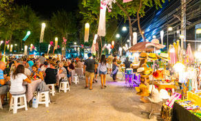 Busy outdoor night market with crowds seated at plastic stools, food stalls and colourful lanterns under palm trees