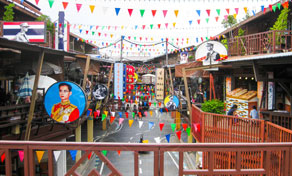 Wooden riverside market walkway in Thailand hung with colourful bunting, a Thai flag and a framed royal portrait