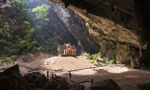 Golden Thai pavilion lit by sunbeam inside vast Phraya Nakhon cave with jungle-covered rock walls