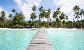 Wooden pier stretching over clear turquoise water toward a palm-fringed white sand tropical beach under blue sky