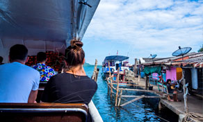 Passengers seated on a boat deck arriving at a stilt village pier with colourful houses over turquoise water