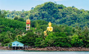 Golden Buddha statue and ornate tower on a jungle hillside above a stilt house at the waterfront
