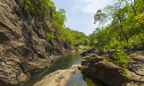 Narrow rocky river gorge with shallow green water, boulders and dense tropical forest under blue sky