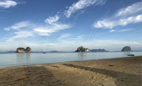 Calm tropical bay with limestone karst islands on the horizon, seen from an empty sandy beach under a cloudy blue sky