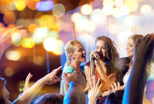 Young women singing into a microphone at a night club, hands raised in crowd against golden bokeh lights