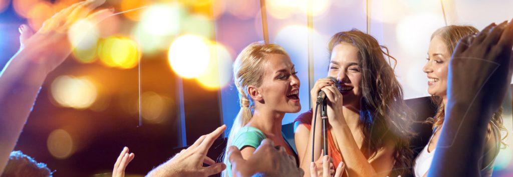 Three young women sing into a microphone at a nightclub, surrounded by raised hands and colourful bokeh lights