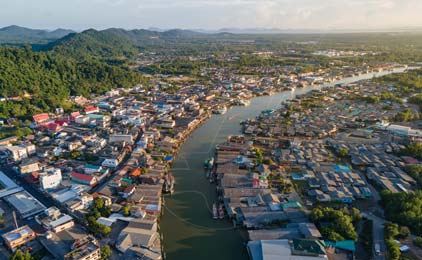Aerial view of riverside town with wooden boats, green hills, and residential buildings along waterway