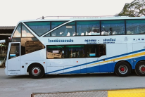 White and blue double-deck coach with Thai lettering on its side, parked on a paved lot beside trees