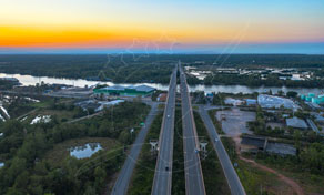 Aerial view at sunset of a divided highway leading to a river bridge, with green woodland and low buildings