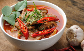 Thai red curry with pork and chilies in a white bowl on a wooden table beside garlic bulbs and dried chili