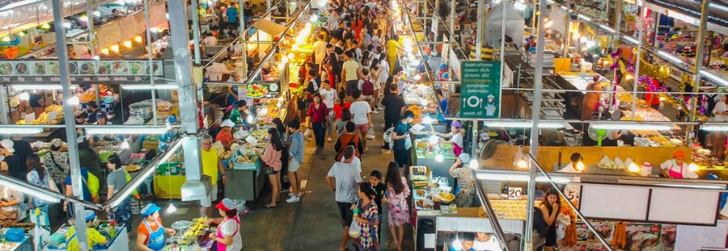 Aerial view of a busy Southeast Asian market with multiple vendor stalls, crowds shopping, and metal framework structure