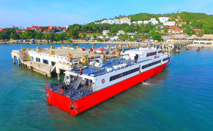 White and red passenger ferry docked at a tropical beach pier with coastal town and green hills in background