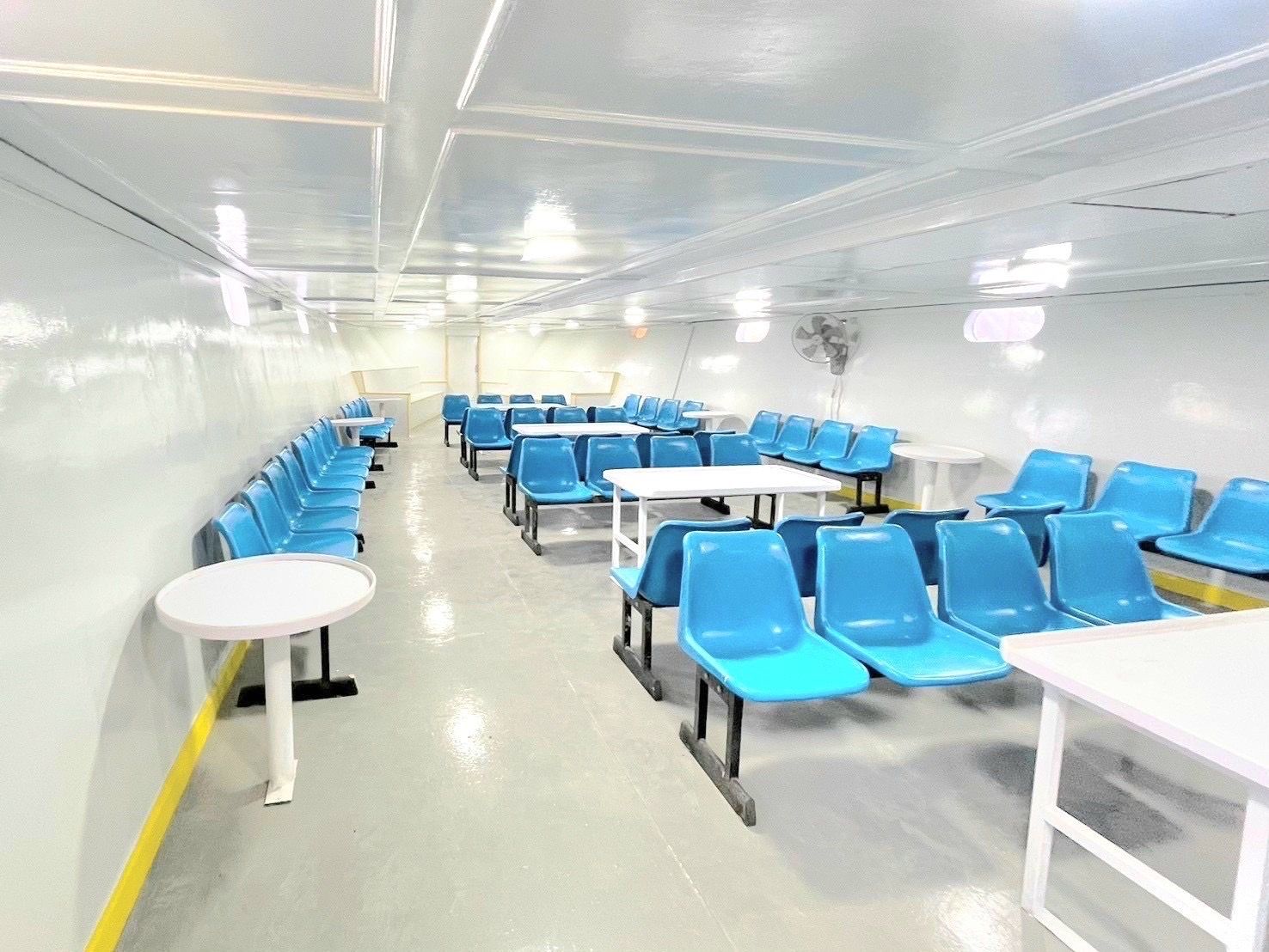 Cabina passeggeri vuota di un traghetto con file di sedili blu in plastica, tavoli bianchi e soffitto bianco illuminato a neon