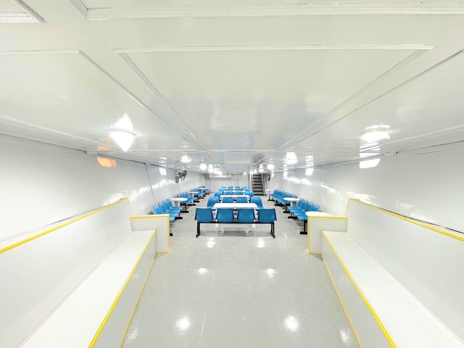 Cabina passeggeri vuota di un traghetto con file di panche blu, tavoli bianchi e pareti e pavimento bianchi lucidi