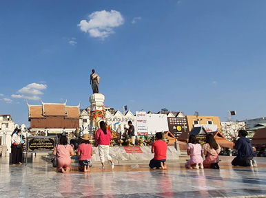 Worshippers kneel and pray before a bronze statue monument in a paved Thai city plaza under blue sky