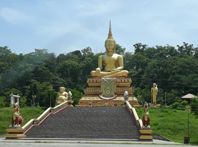 Large golden seated Buddha statue atop a wide stone staircase at a Thai temple, framed by green trees