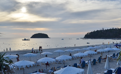 Beach scene with white umbrellas, visitors, boats, and forested islands under cloudy sky at dusk