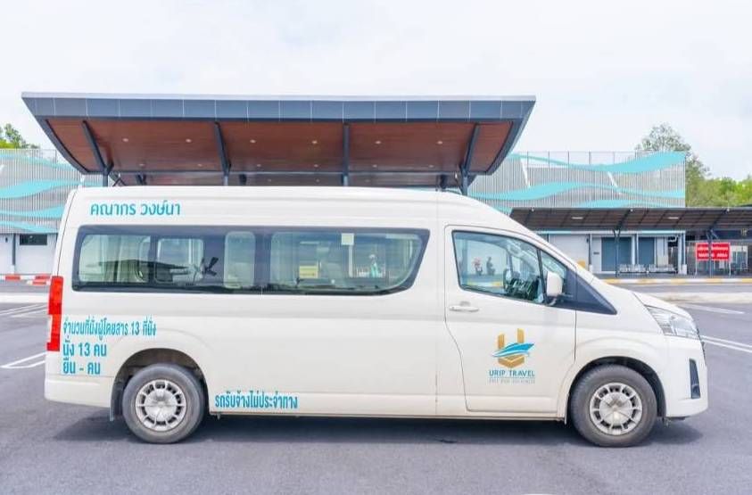 Weißer 13-Sitzer-Minivan mit thailändischer Schrift unter einem überdachten Terminalvordach auf grauem Asphalt