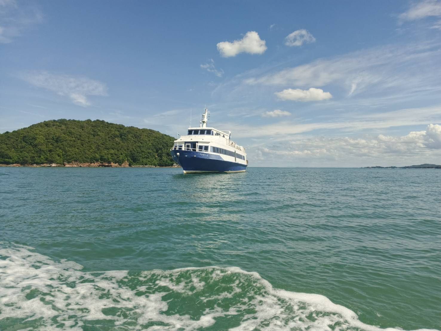 Blau-weiße Passagierfähre vor Anker im türkisen Meer neben grün bewaldeter Insel unter blauem Himmel