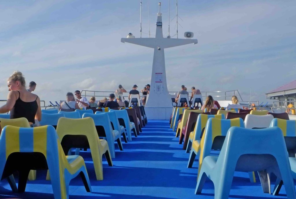 Open-air ferry deck with blue and yellow seating, passengers, and structural framework against cloudy sky