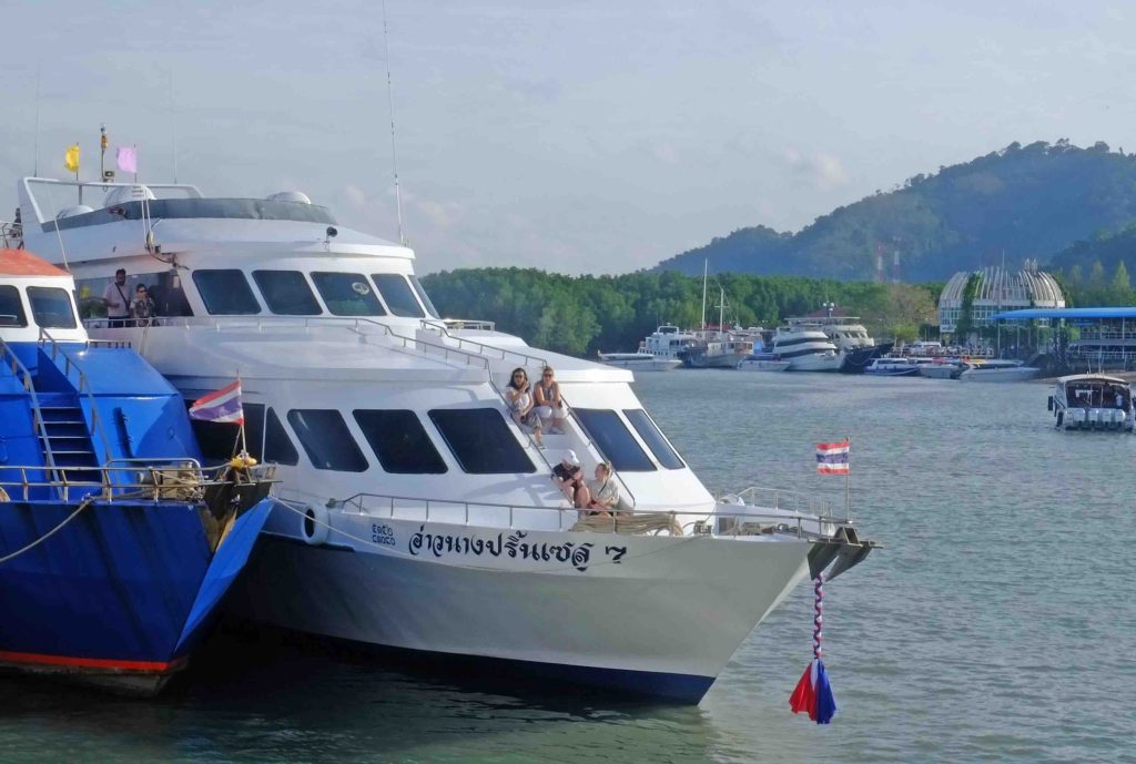White and blue passenger ferry docked at pier with tourists boarding, mountainous coastline in background