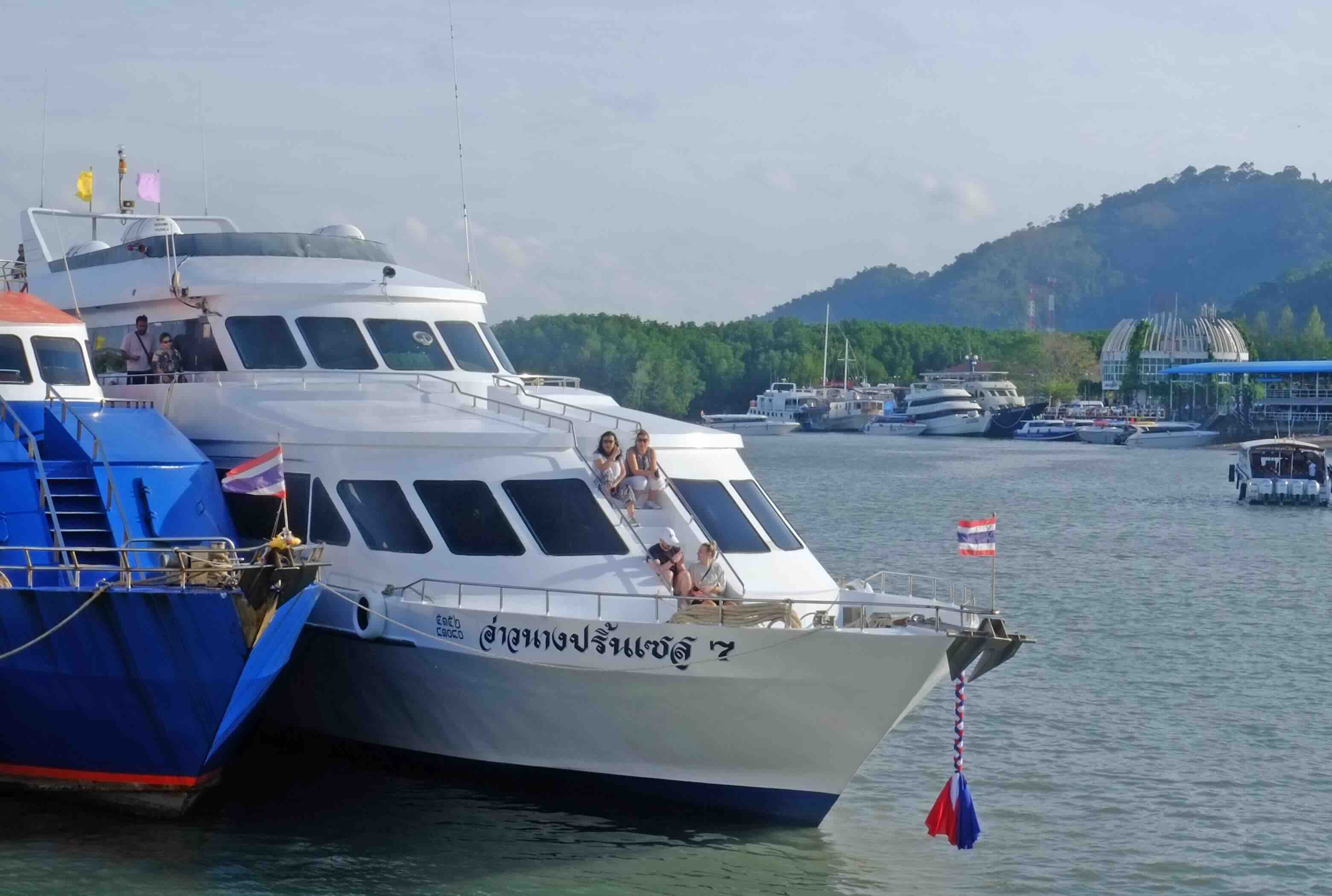 White and blue passenger ferry docked at pier with tourists boarding, mountainous coastline in background
