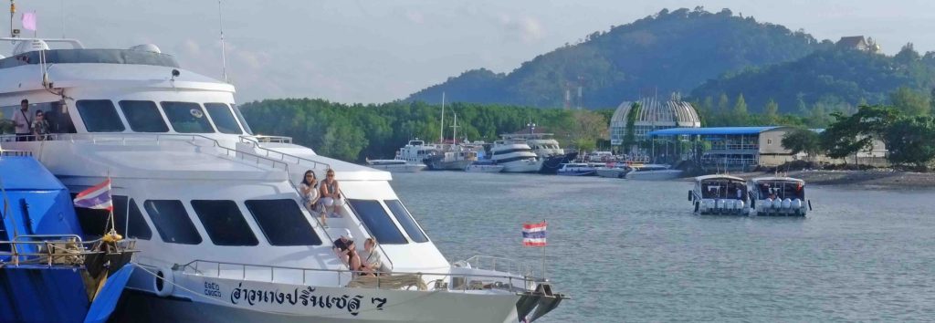 White passenger ferry with tourists at a busy Southeast Asian pier, forested hillside and moored boats in background