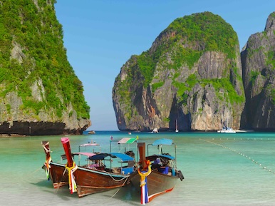 Longtail boats moored on turquoise shallows below towering limestone cliffs, Maya Bay-style Thai island beach