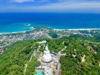 Aerial view of the white Big Buddha statue on a forested Phuket hilltop above turquoise sea and coastal town