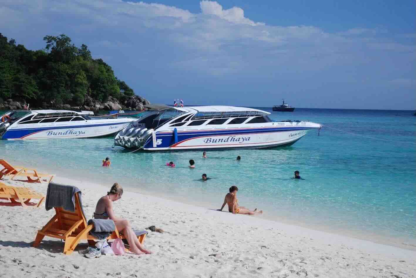 Speedboote mit Aufschrift „Bundhaya“ am weißen Sandstrand, Badende im klaren türkisen Wasser