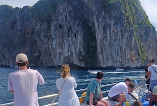 Tourists on a boat viewing a tall limestone cliff rising from blue water