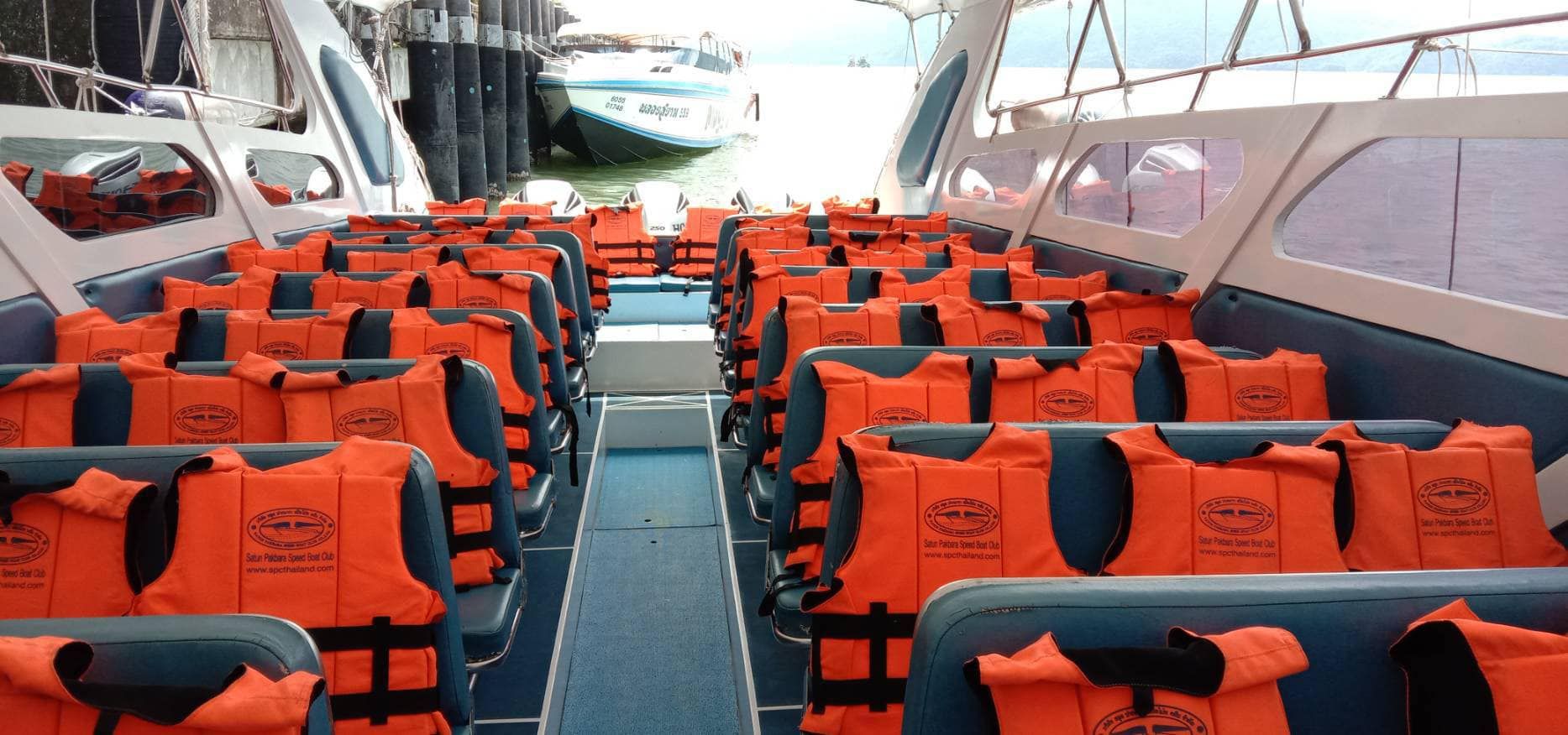 Speedboot-Passagierkabine mit blauen Sitzreihen voller orangefarbener Rettungswesten, dahinter Boot am Pier