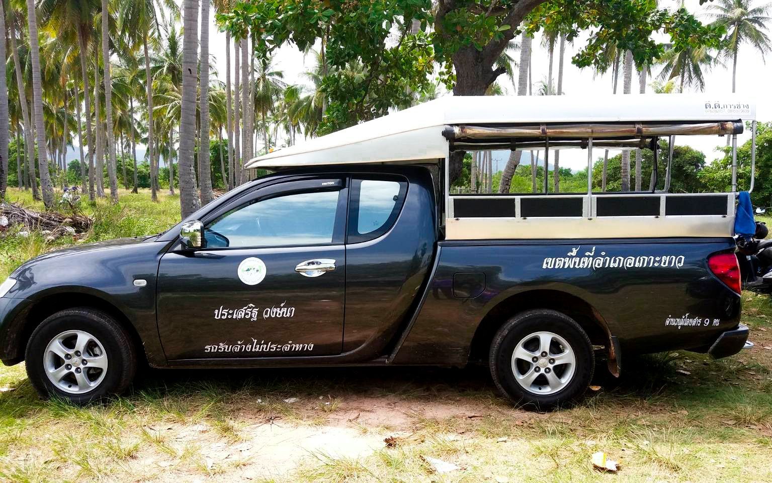 Dunkelgrauer Pickup, umgebaut zum thailändischen Songthaew-Taxi mit Dachverdeck, unter Kokospalmen geparkt