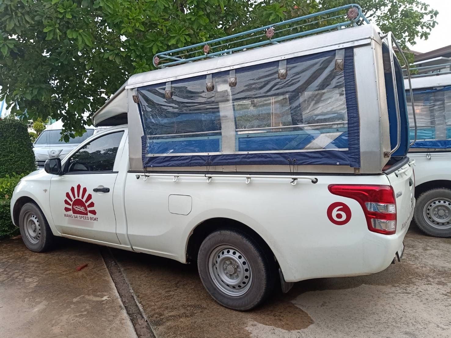 Weißer Songthaew-Pickup mit überdachter Sitzbank, rotem Sonnenlogo für Bootstransfers und Nummer 6 auf Beton