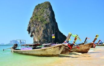 Wooden longtail boats moored on a turquoise shallow beach below a towering limestone karst, Railay-style coast, Thailand