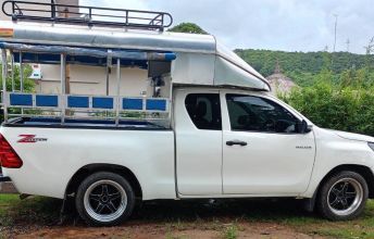White pickup truck converted to a songthaew with blue bench seats and roof rack parked on grass by green hills