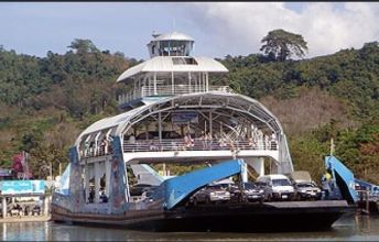 Multi-deck car ferry with open bow ramp and vehicles on deck moored below forested green hills