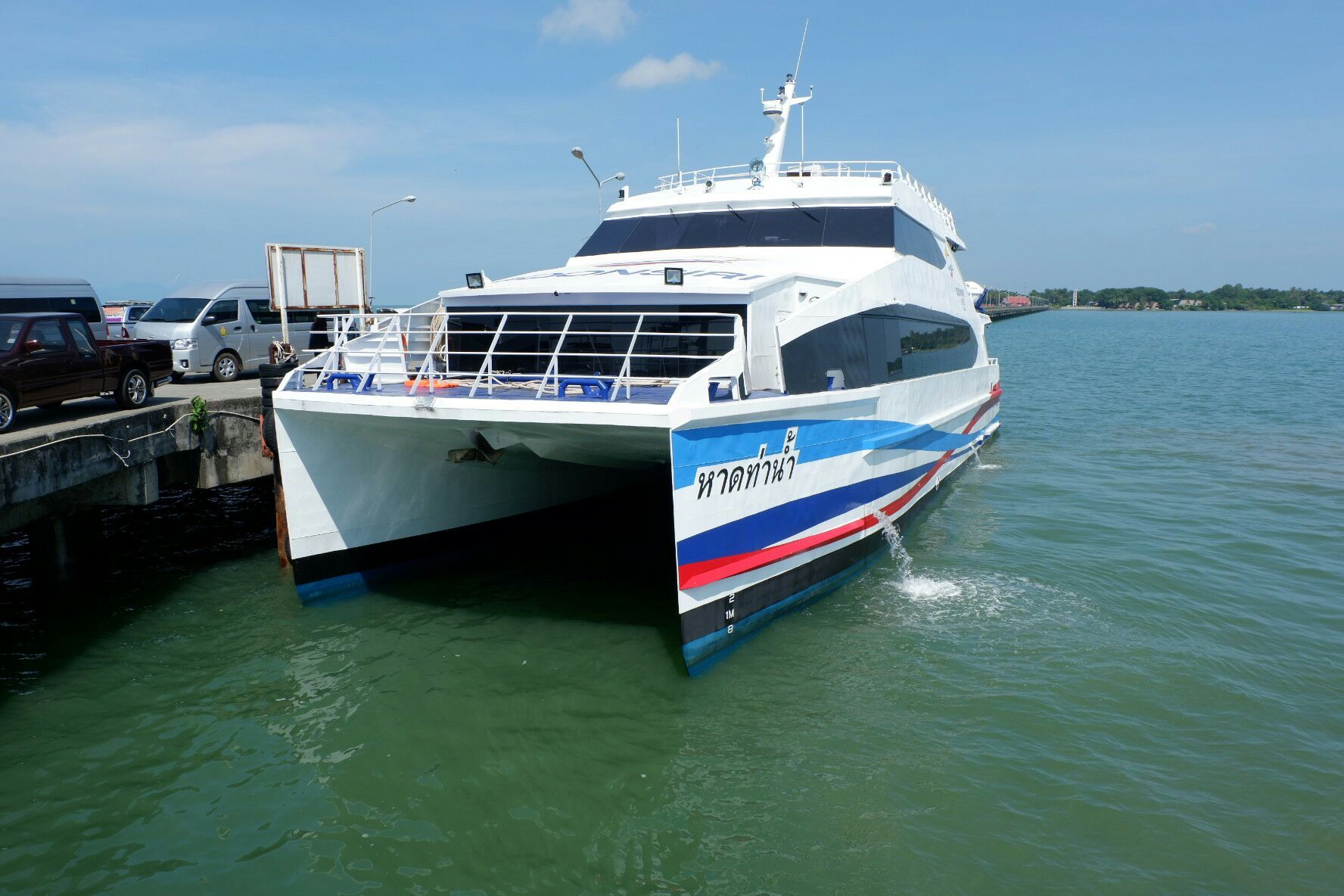 Weiße Hochgeschwindigkeits-Katamaranfähre mit Thai-Schrift am Rumpf an einem Betonpier unter blauem Himmel