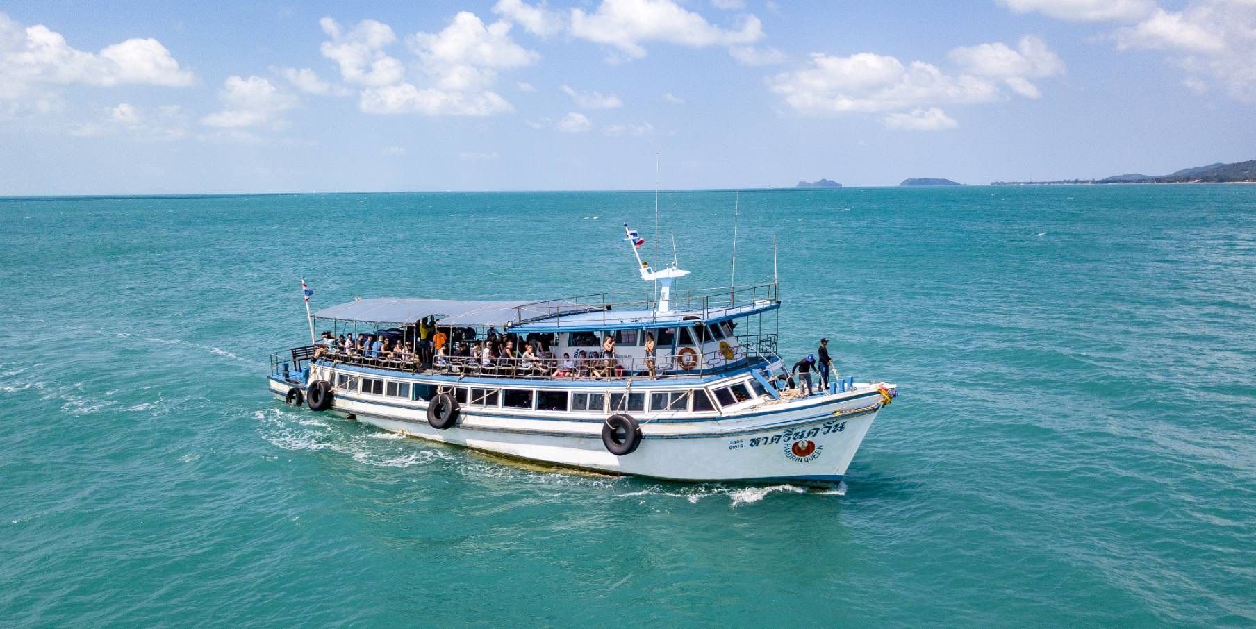 Luftaufnahme einer hölzernen Thai-Passagierfähre voller Touristen auf türkisem Meer, Inseln am Horizont
