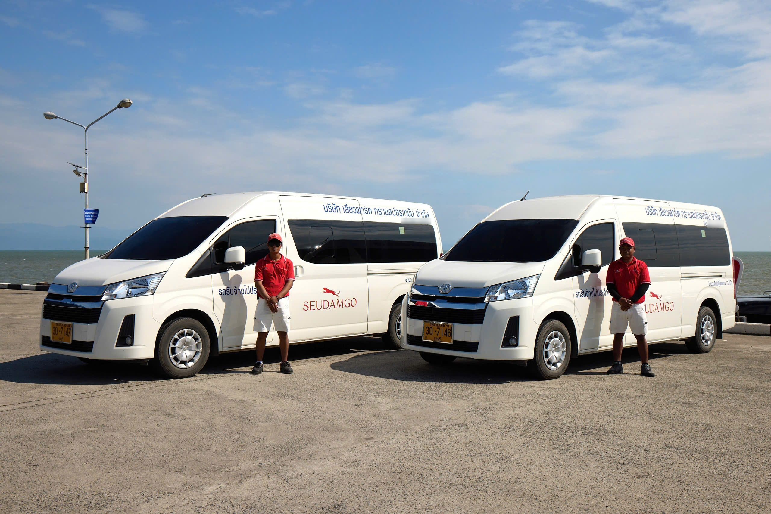 Two white Toyota minivans with red company branding parked on a concrete pier by the sea, drivers in red shirts standing beside
