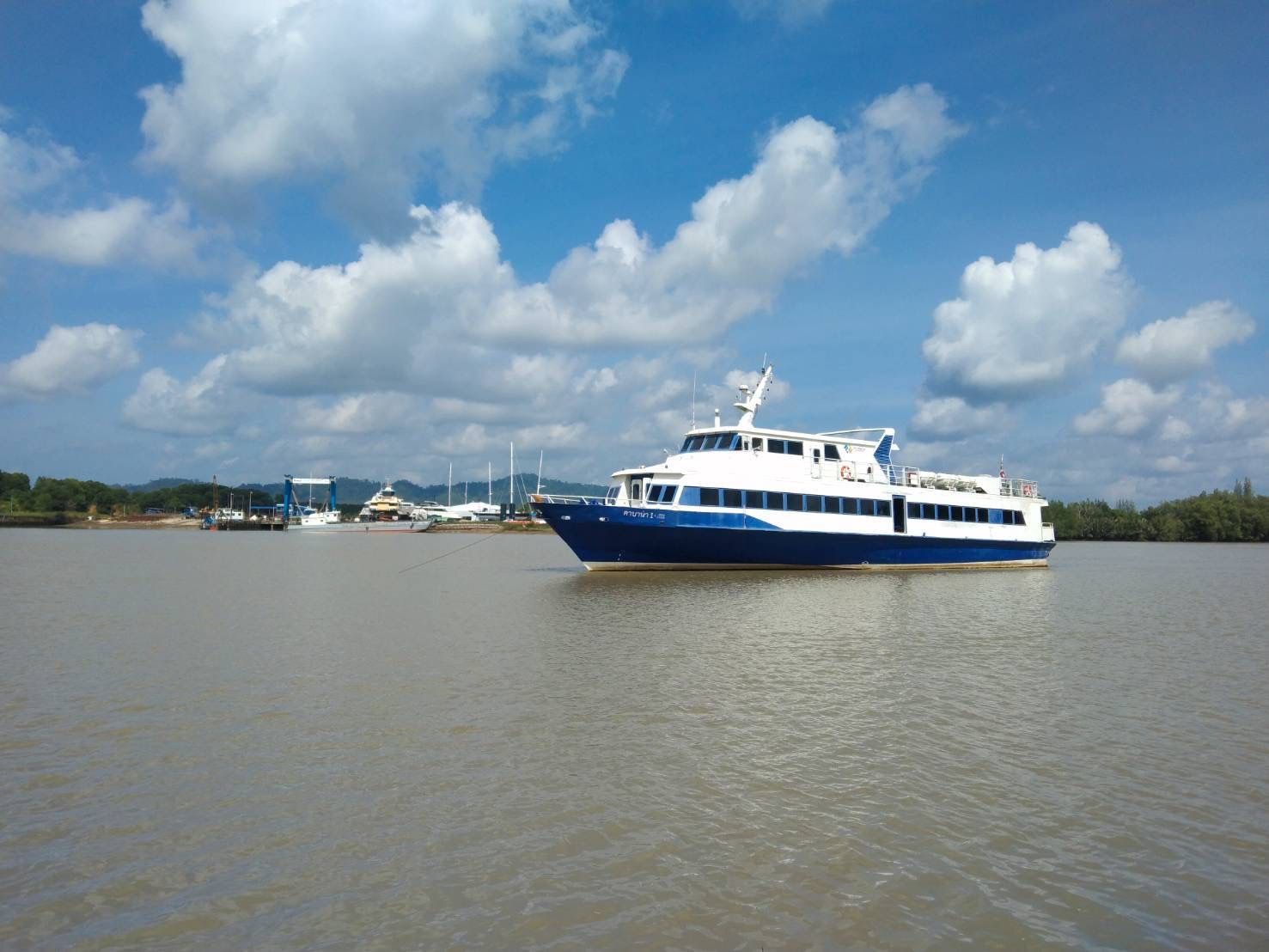 Blau-weiße Passagierfähre ankert auf braunem Gezeitenfluss neben Mangroven, dahinter ein kleiner Yachthafen